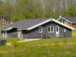 A modern house with a dark exterior, white trim, and a sloped roof. It features a front porch with a bench and a small garden area with yellow flowers. - 4 Star Holiday Home in Sydals (Sydals)