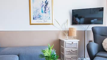Una sala de estar con un sofá azul, una mesita auxiliar blanca con una planta, una obra de arte enmarcada y un televisor en la pared.
