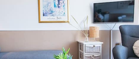 A living room with a blue sofa, a white side table with a plant, a framed artwork, and a television on the wall.