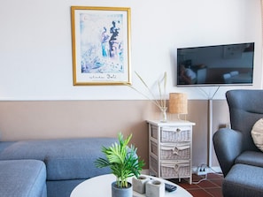 A living room with a blue sofa, a white side table with a plant, a framed artwork, and a television on the wall.