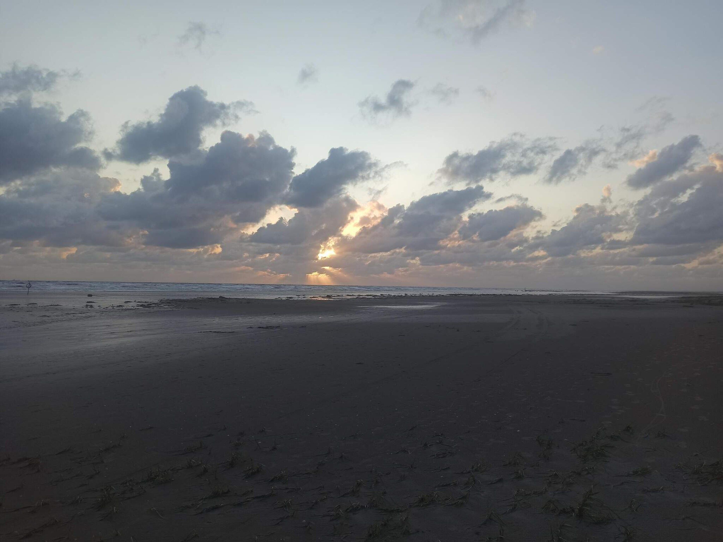 Uma praia com céu nublado e o sol se pondo ou nascendo.