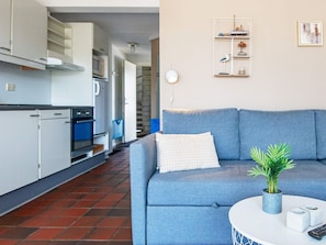 Un salon moderne avec un canapé bleu, une petite table ronde agrémentée d’une plante, et un coin cuisine doté de meubles blancs et d’une cuisinière.