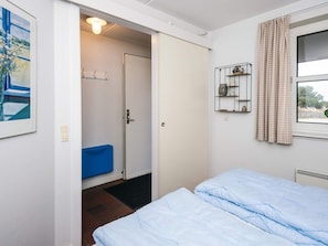 A bedroom with a bed, a door leading to a bathroom, a shelf with decorative items, and a window with curtains.