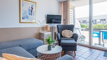 Una sala de estar moderna con un sofá, un sillón y una mesita con una planta. Un televisor está montado en la pared, y hay una ventana con cortinas.