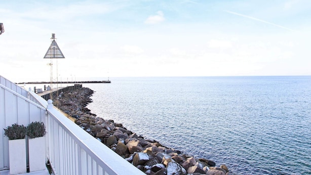 A coastal view with a white building, a rocky breakwater, and a pier extending into the sea. - Seaside Escape in Bagenkop - By Traum Ferienwohnungen (Sydlangeland)