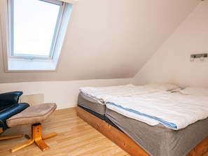 A small, single-bed room with a sloped ceiling, a window, a desk with a chair, and a wall-mounted light fixture.