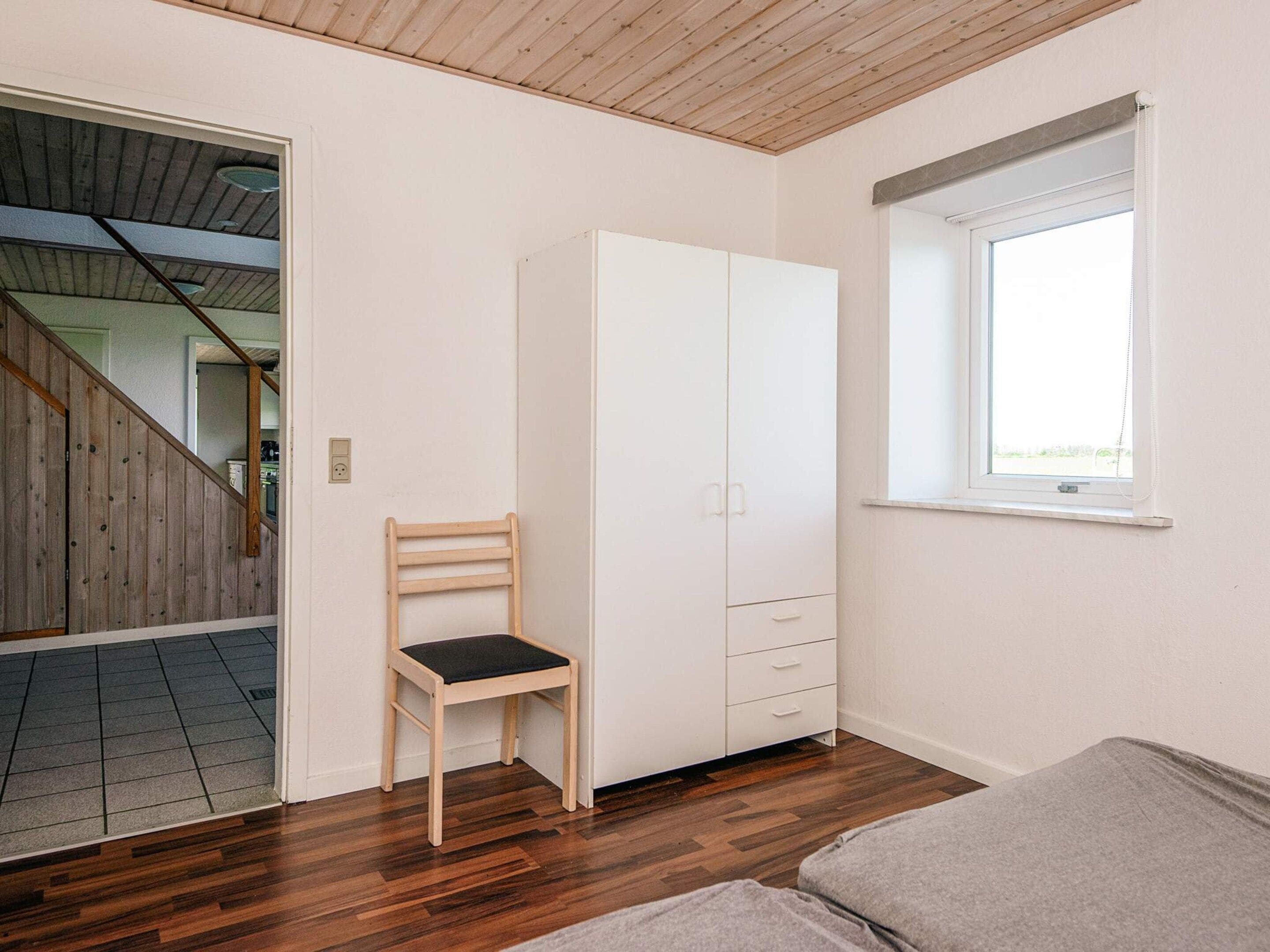Una habitación con piso de madera, un armario blanco, una silla de madera y una ventana con vistas.