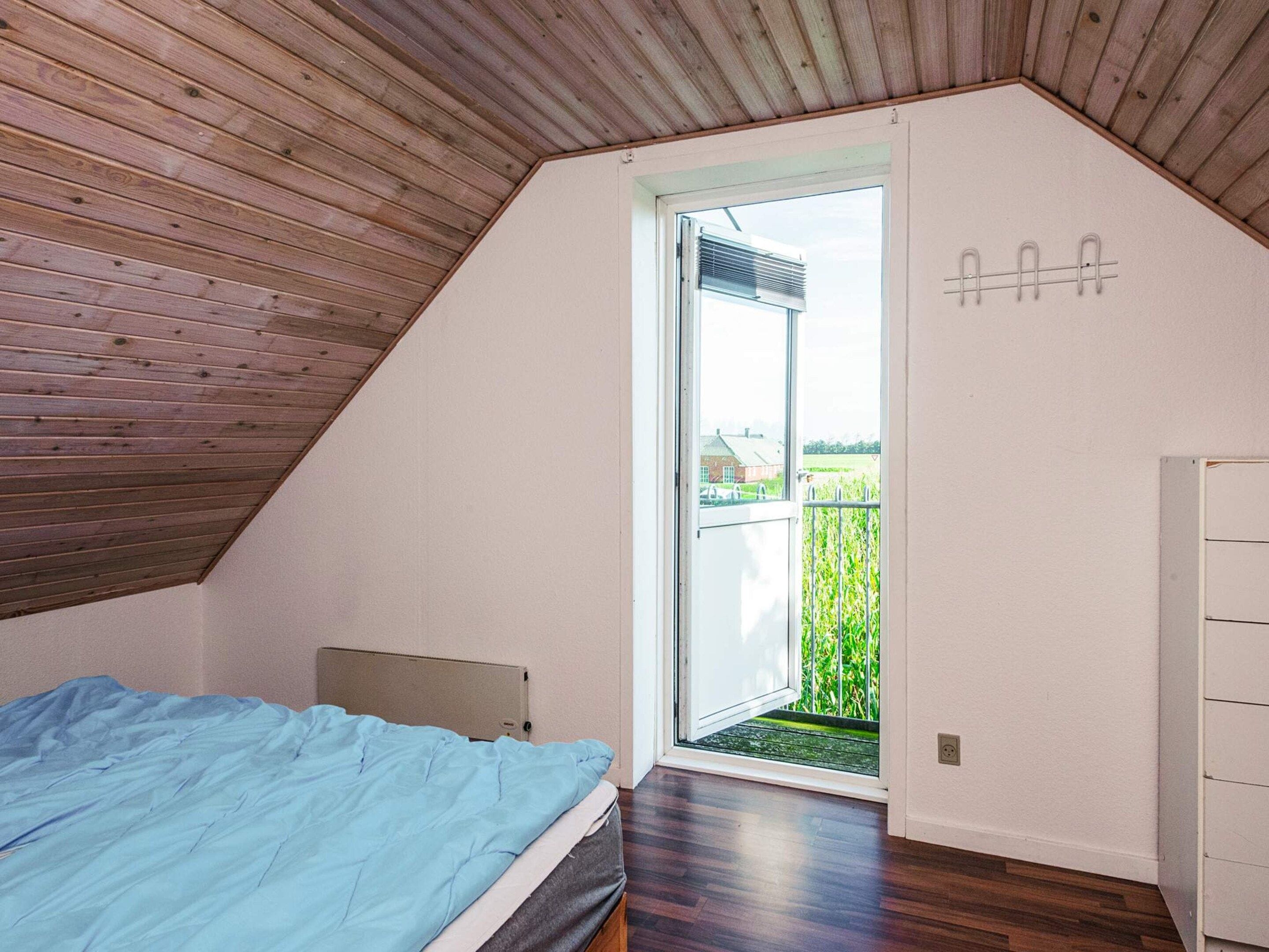 Ein Schlafzimmer mit schrägen Wänden, einem Bett mit blauer Bettdecke, einer Tür zu einem Balkon und einer weißen Wand mit einem Metallregal.