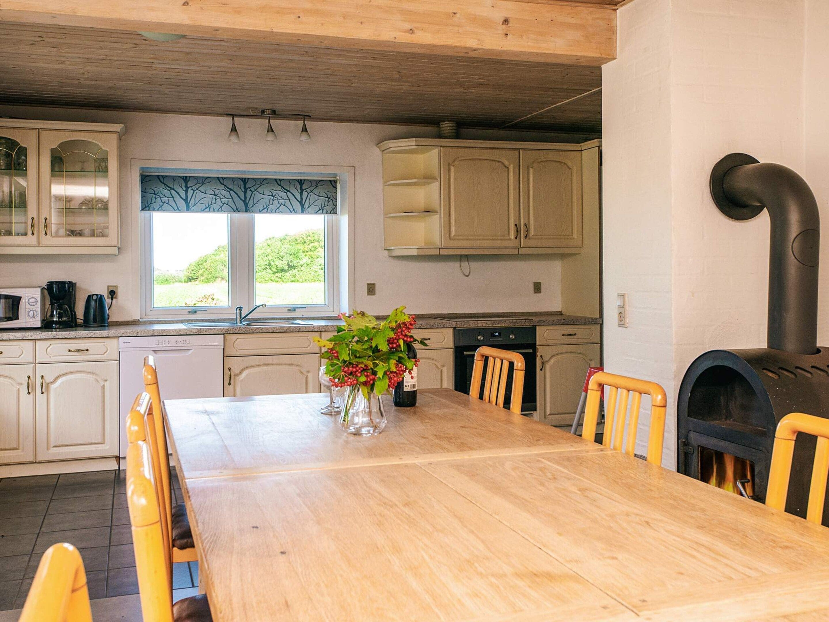 Une cuisine avec des meubles en bois, une table à manger, des chaises, une fenêtre donnant sur un beau paysage et un poêle à bois.