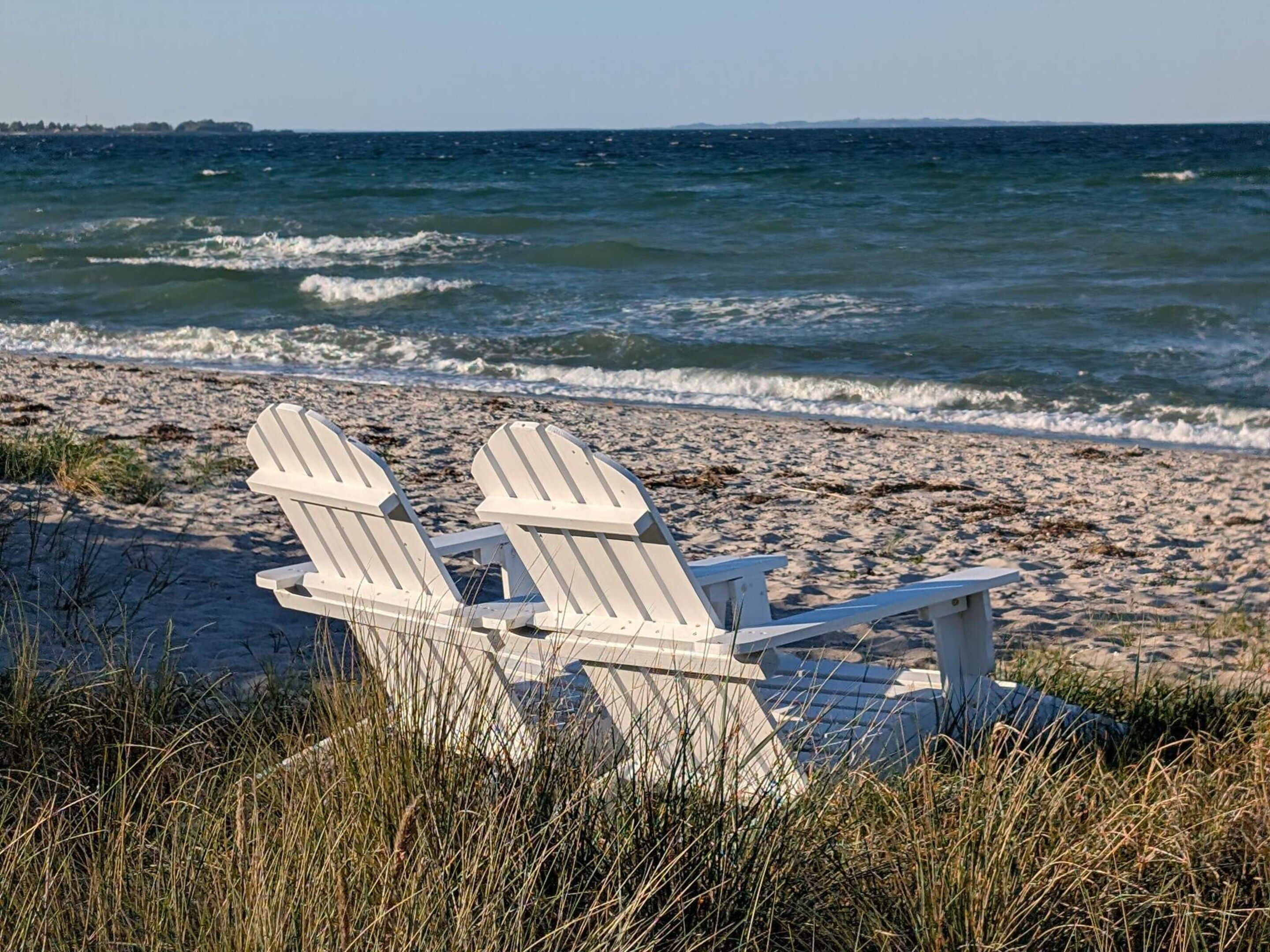 Zwei weiße Adirondack-Stühle an einem sandigen Strand, im Hintergrund der Ozean.