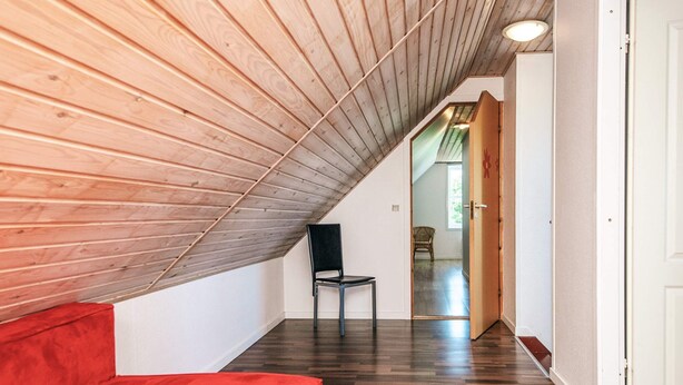 A room with a sloped wooden ceiling, a red sofa, a black chair, and a wooden floor. - 12 Person Holiday Home in Bredebro (Bredebro)