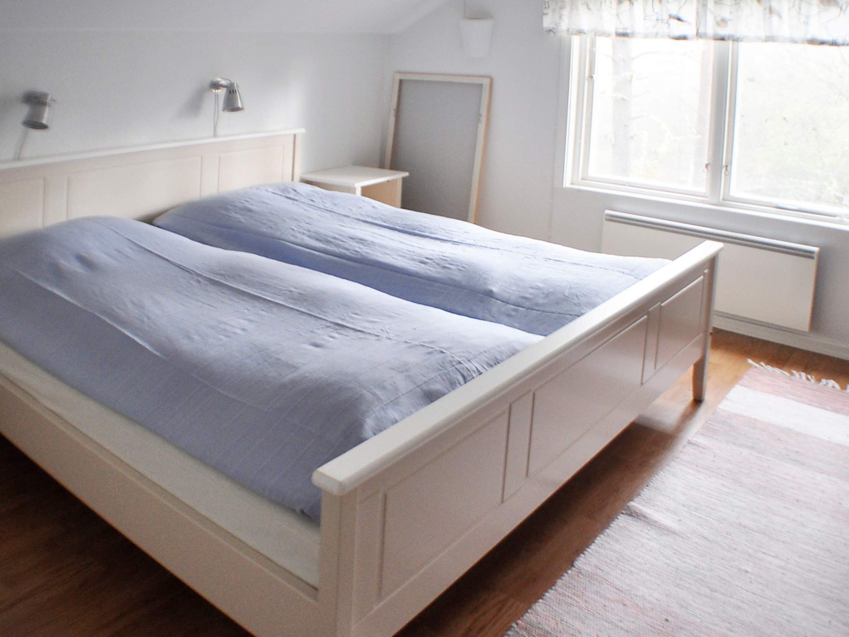 Cama doble con estructura blanca y cubrecama azul en habitación con piso de madera, mesita de noche pequeña, espejo y ventana con cortinas.