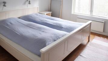Cama doble con estructura blanca y cubrecama azul en habitación con piso de madera, mesita de noche pequeña, espejo y ventana con cortinas.