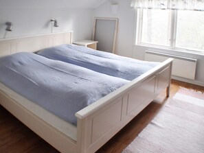 Un letto matrimoniale con struttura bianca e copripiumino blu in una stanza con pavimento in legno, un comodino, uno specchio e una finestra con tende.
