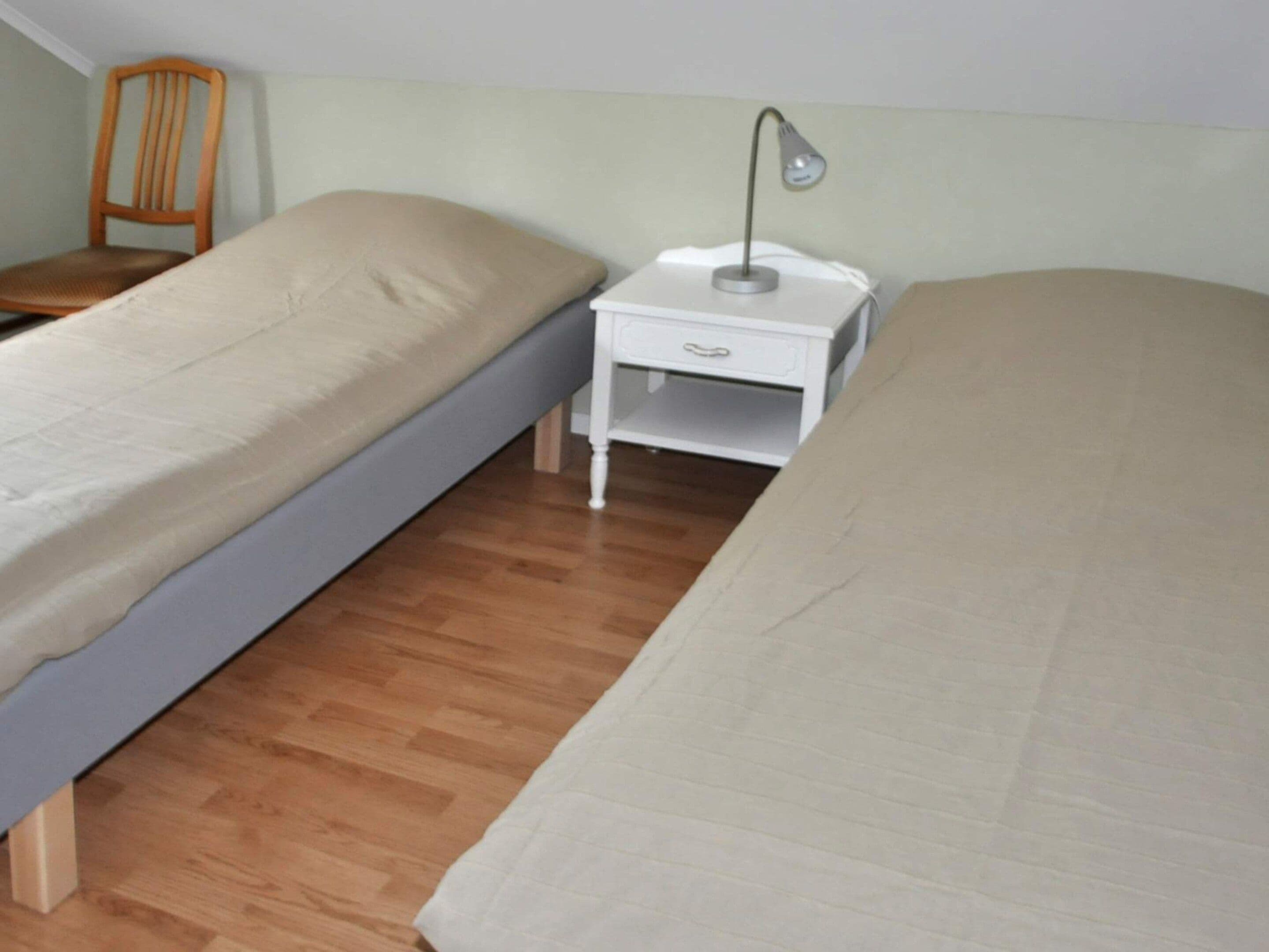 A small, single-room with two single beds, a wooden chair, a white bedside table with a lamp, and a wooden floor.