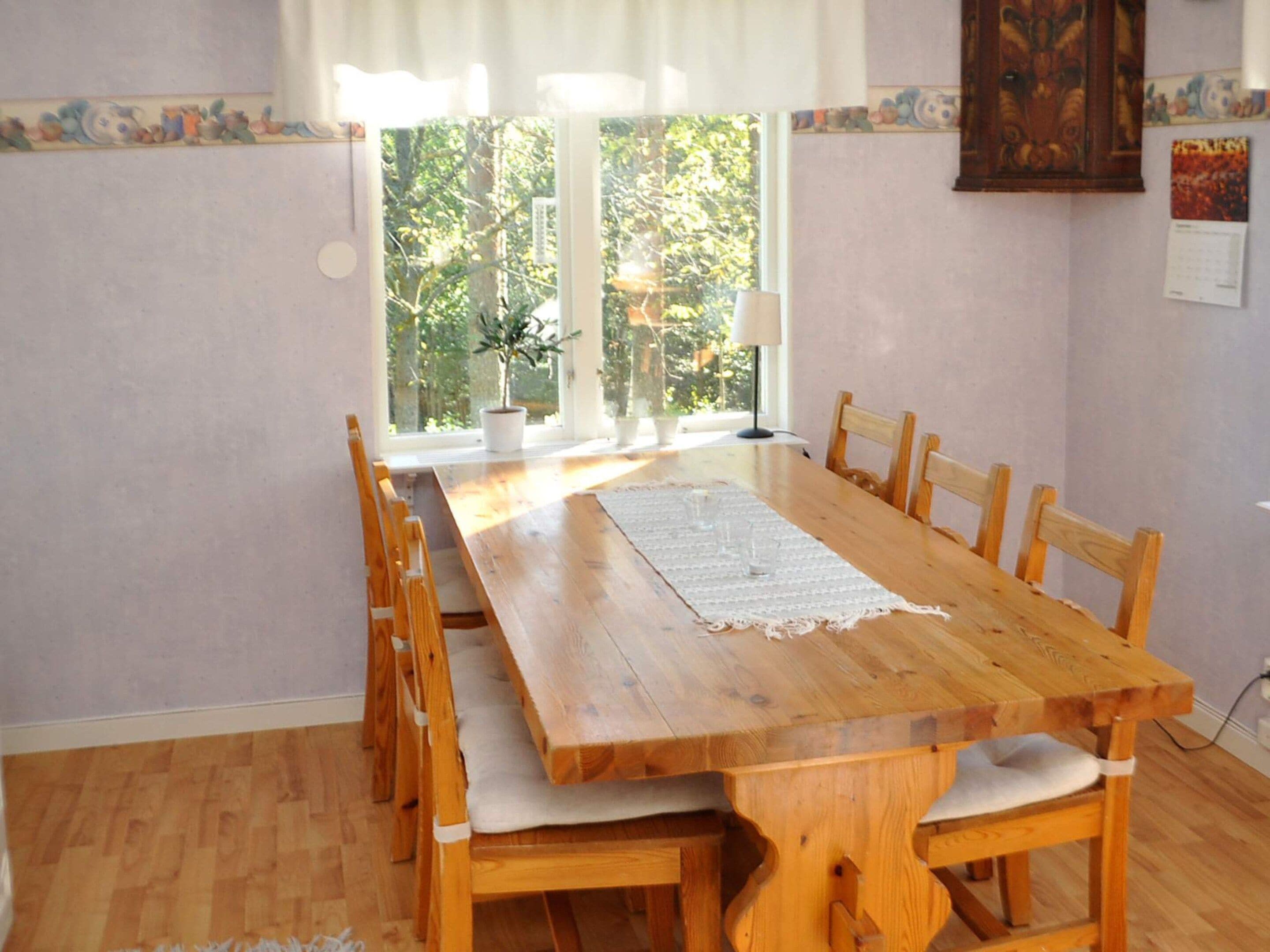 Uma área de refeições com mesa e cadeiras de madeira, um jogo de mesa de renda e uma janela com vista para vegetação.