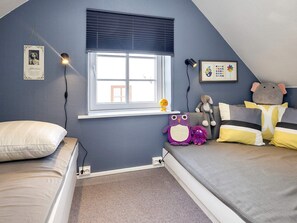 Quarto com teto inclinado, janela com persianas, mesa de cabeceira, luminária e prateleira na parede com brinquedos de pelúcia.