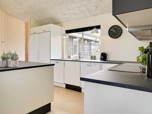 A modern kitchen with white cabinets, a black countertop, a clock, and a window with blinds. - 5 Star Holiday Home in Bogense (Bogense)