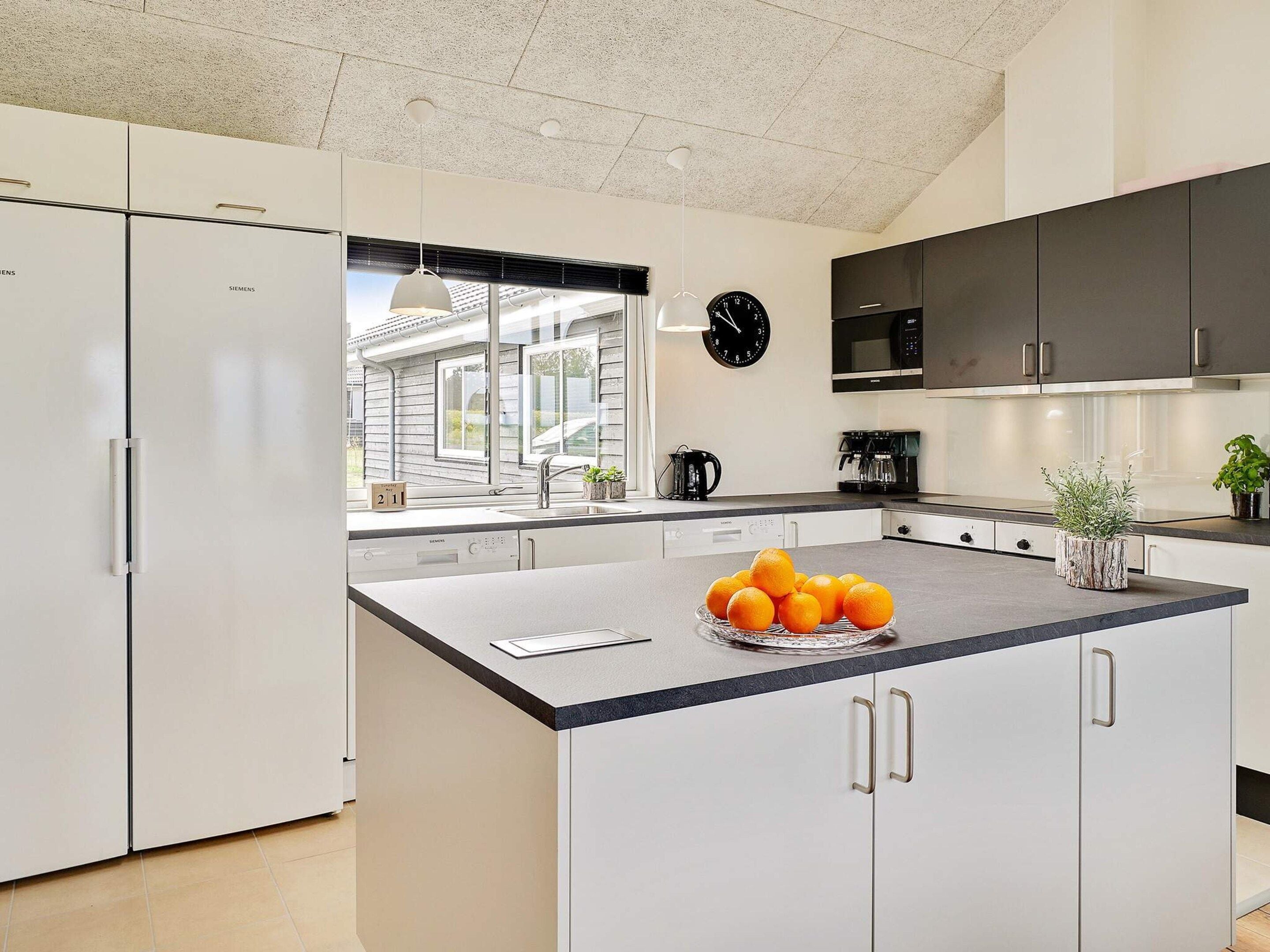 Cocina moderna con armarios blancos, una isla central con frutero, un reloj negro y una ventana con persianas.