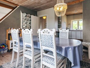 A dining area with a round table, white chairs with patterned upholstery, a chandelier, and a view of the kitchen through an open door.