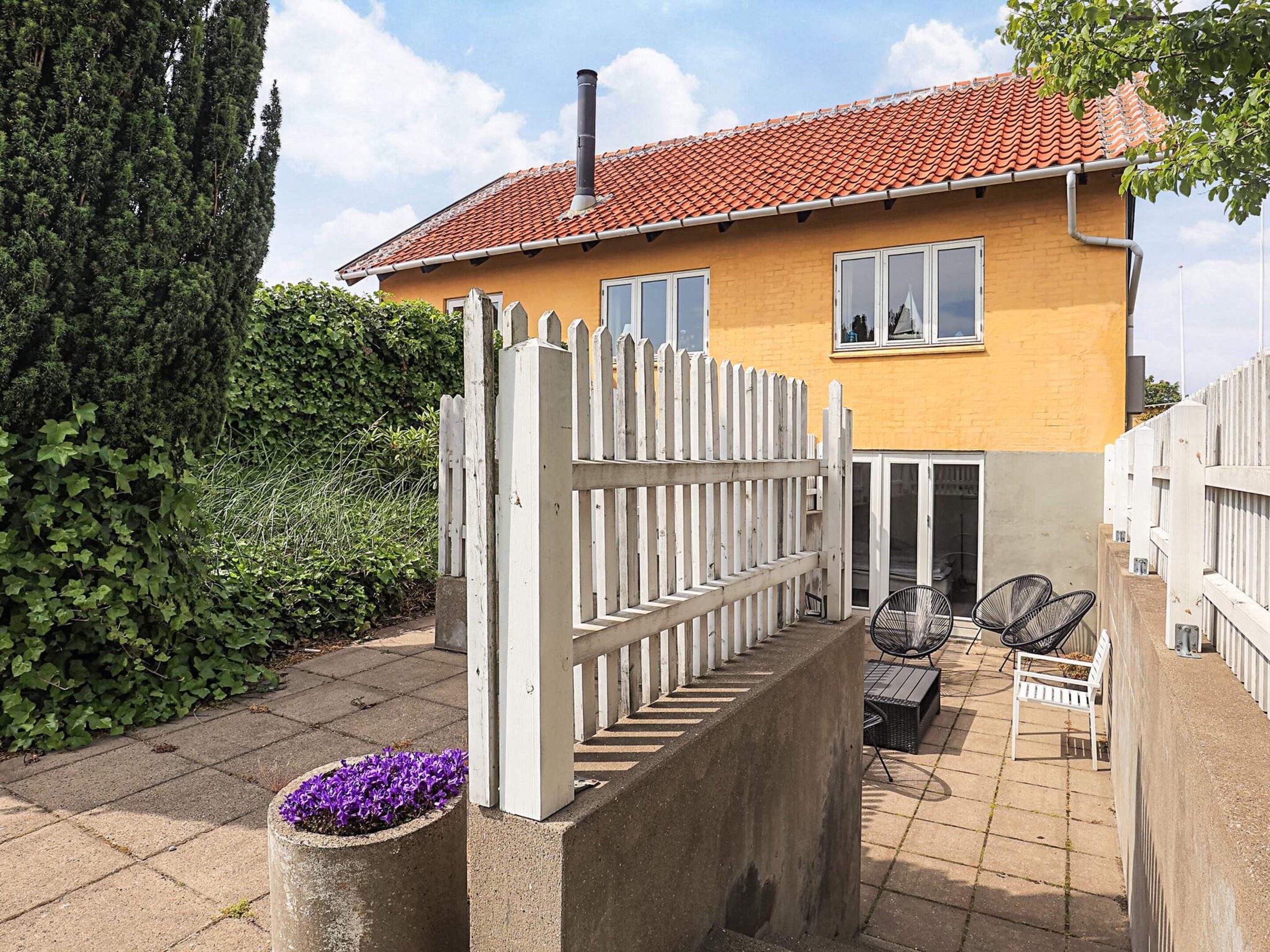 A two-story house with a red-tiled roof, a white picket fence, and a patio area with outdoor furniture.