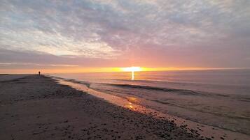 A beach with a person walking, the sun setting in the distance, and the ocean.