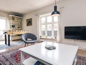 Una sala de estar moderna con una mesa blanca, un sillón gris, una lámpara colgante negra, un televisor y una ventana con cortinas.