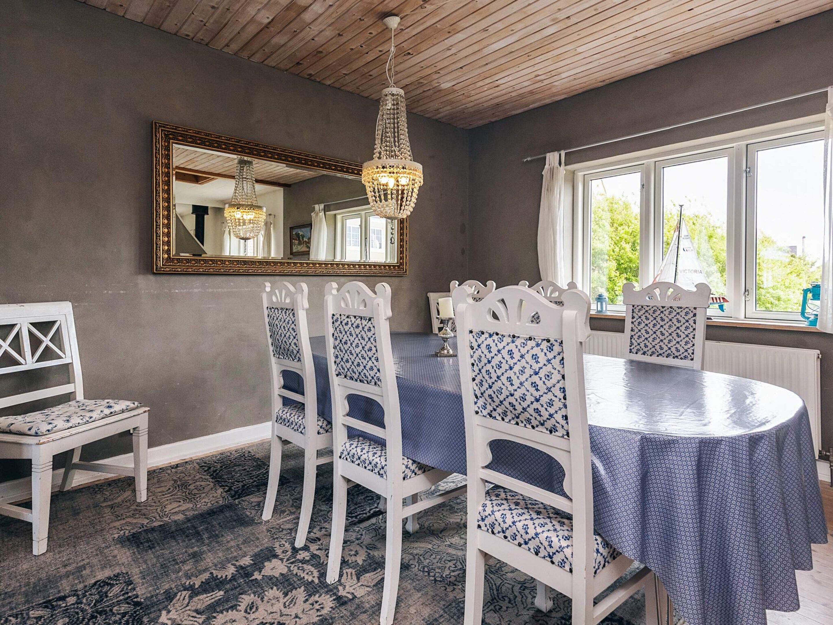 A dining area with a round table, white chairs, a patterned rug, a mirror, and a window with a view of greenery.