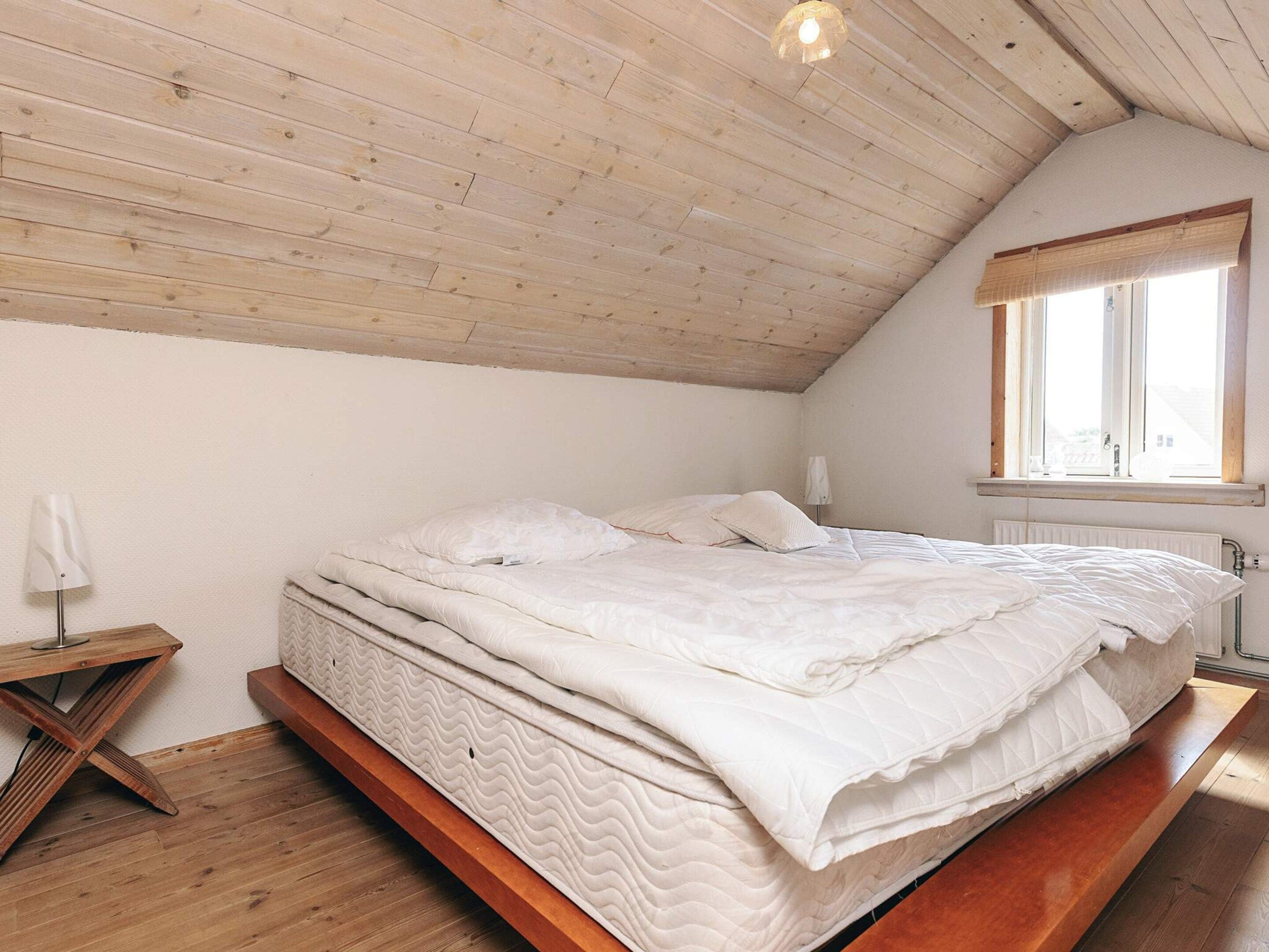 Een slaapkamer met een schuine kap, een bed met wit beddengoed, een nachtkastje, een lamp en een raam met uitzicht.