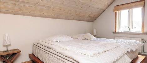 Een slaapkamer met een schuine kap, een bed met wit beddengoed, een nachtkastje, een lamp en een raam met uitzicht.
