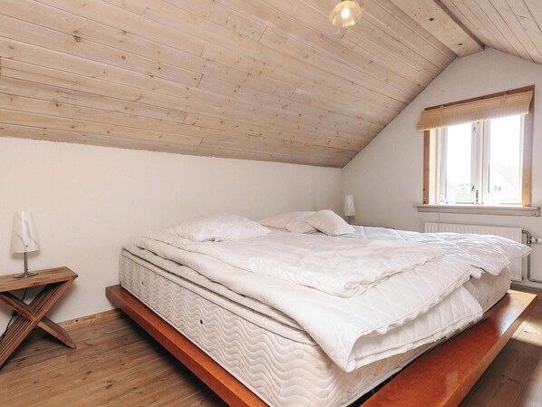 Un dormitorio con techo inclinado, una cama con ropa de cama blanca, una mesita de noche, una lámpara y una ventana con vistas.