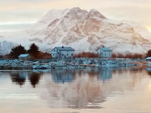 A mountain landscape with a reflection in a calm body of water, featuring two houses with a snowy foreground. - 5 Person Holiday Home in Vandve (Dønna)