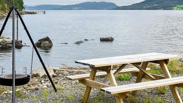 A wooden picnic table by a calm lake with a tripod and a grill. - 7 Person Holiday Home in Averoy (Averoy)