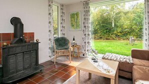 A cozy living room with a fireplace, a comfortable chair, a table, and a view of a lush garden through large windows. - Family Fun in Ristinge - By Traum Ferienwohnungen (Sydlangeland)