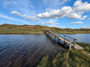 A wooden bridge over a calm body of water with grassy banks and hills in the background.