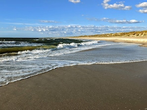 Uma praia com ondas suaves quebrando na costa, céu claro e paisagem montanhosa ao longe.