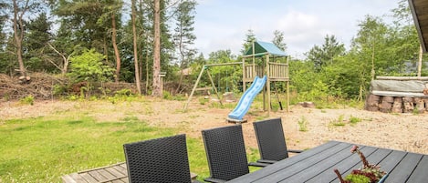 A wooden deck with a table and chairs, a play structure with a slide, and a garden with various plants.