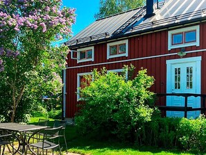 A red building with white-framed windows and a door, surrounded by lush greenery and a tree with purple flowers.