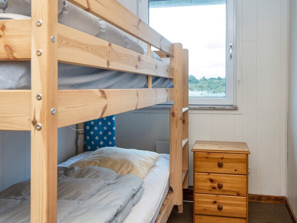 A bunk bed with a wooden frame, a bedside table with drawers, and a window with a view of greenery.