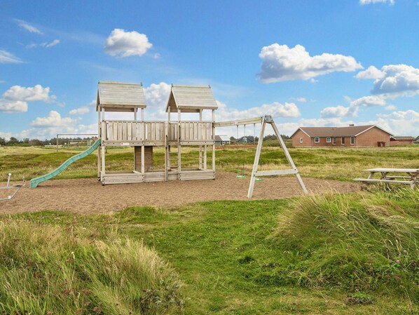 Une aire de jeux avec deux cabanes en bois, un toboggan et des balançoires.