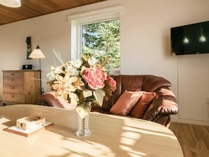 Una habitación con una mesa de madera, un jarrón con flores, un sofá de cuero marrón, un armario de madera y una ventana con vistas a árboles.