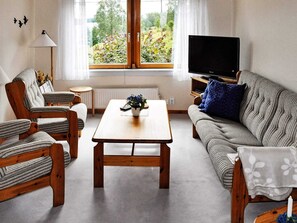 Een woonkamer met een bank, stoelen, een salontafel, een televisie en een raam met uitzicht op groen.