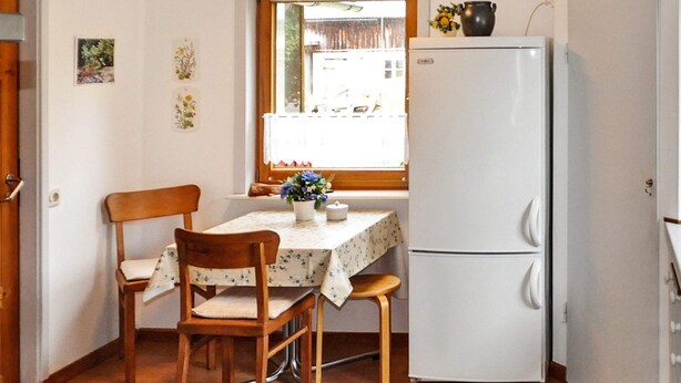 Eine kleine Küche mit einem weißen Kühlschrank, Holzstühlen, einem Tisch mit Blumenmuster und einem Fenster mit Blick nach draußen.