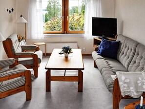 A living room with a wooden coffee table, striped armchairs, a sofa with a blue pillow, a television on a wooden stand, and a window with a view of greenery. - 6 Person Holiday Home in Bengtsfors (Bengtsfors)