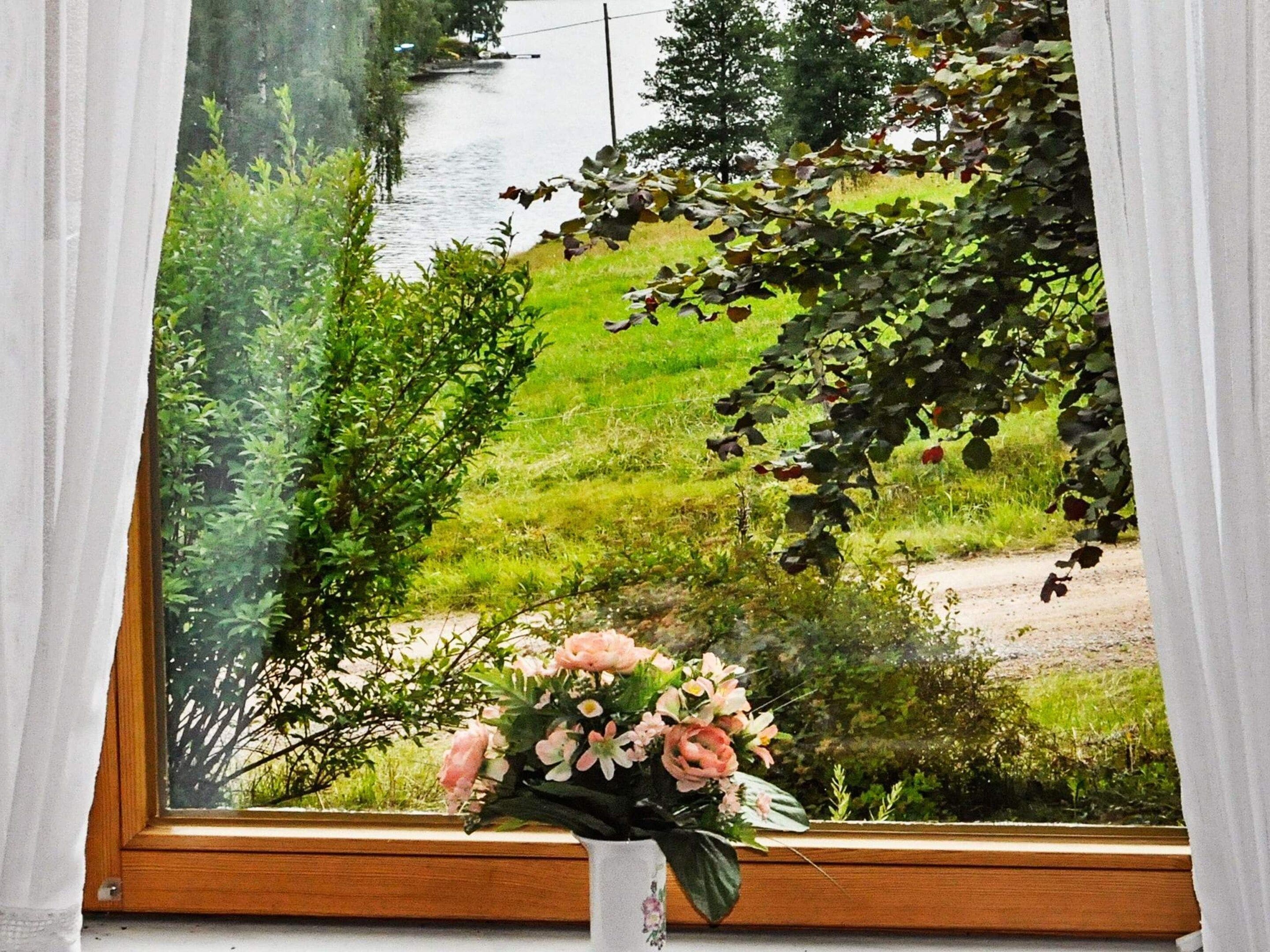 A window with a view of a river and greenery, a wooden frame, and a bouquet of flowers on the windowsill.