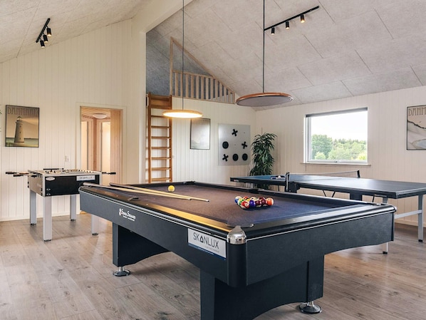 A pool table with a ball rack, a foosball table, and a billiards table in a room with wooden flooring and white walls.
