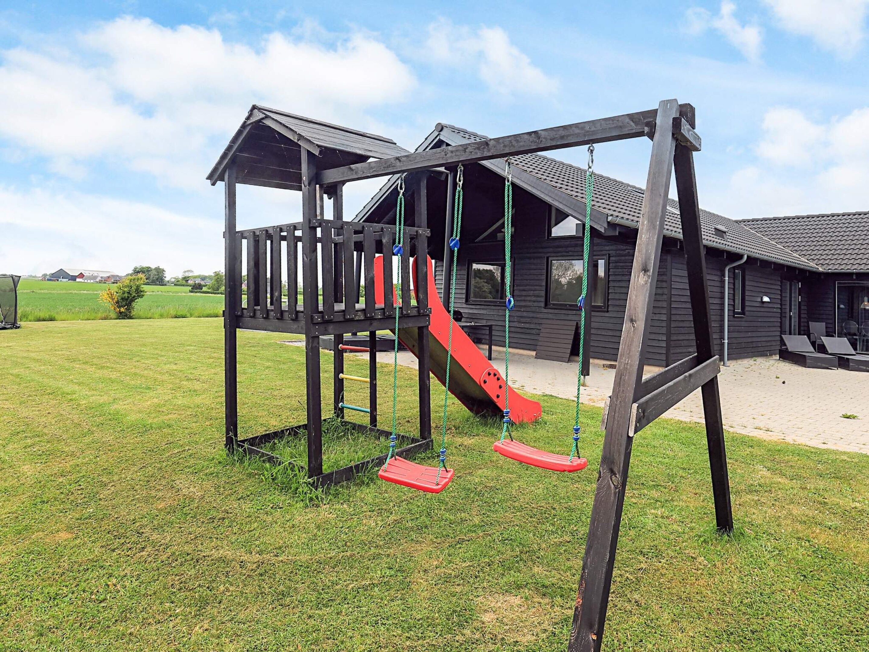 A playground with a wooden structure, including a slide and swings, in front of a building with a tiled roof.