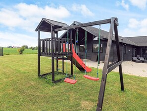 Een speeltuin met een houten constructie, inclusief een glijbaan en schommels, voor een gebouw met een pannendak.