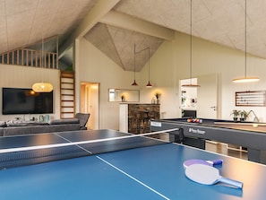 Una mesa de ping pong con red, un sofá, un televisor y una zona de bar con taburetes.