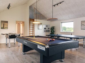 Una mesa de billar con un soporte para las bolas, una mesa de fútbol de mesa y una mesa de carambola en una habitación con pisos de madera, paredes blancas y una ventana.